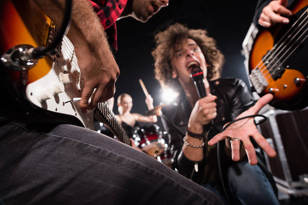 KYIV, UCRANIA - 25 de agosto de 2020: Vista de bajo ángulo del vocalista gritando en micrófono cerca de guitarristas con retroiluminación sobre fondo borroso - Foto, Imagen