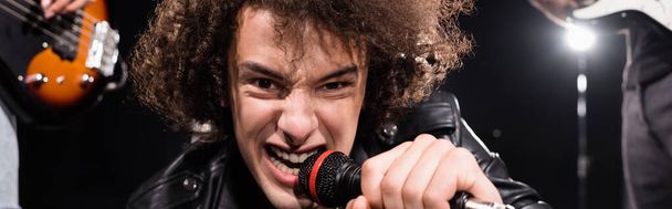 KYIV, UKRAINE - AUGUST 25, 2020: Angry vocalist with microphone signing while looking at camera with backlit and blurred guitarists on black, banner - Foto, Imagen