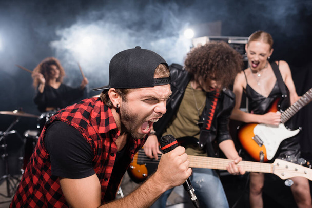 KYIV, UKRAINE - AUGUST 25, 2020: Vocalist screaming in microphone while leaning forward near rock band musicians on blurred background - Foto, Imagen