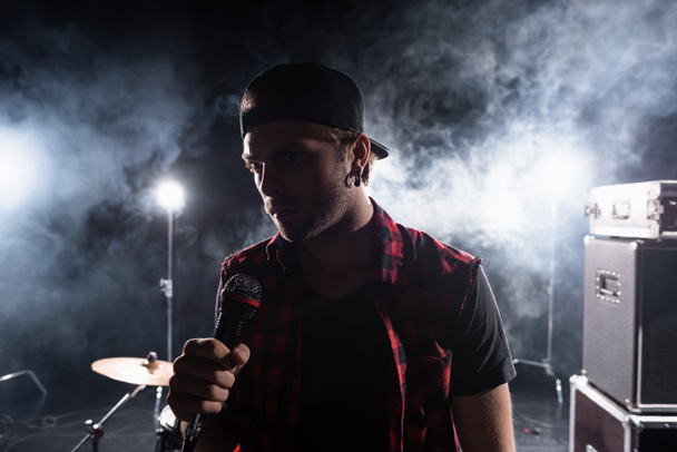 Vocalista de banda de rock sosteniendo micrófono, con humo y retroiluminado sobre fondo borroso - Foto, Imagen