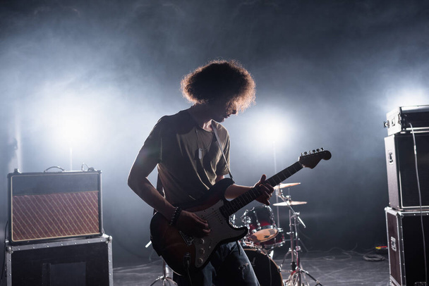 KYIV, UKRAINE - AUGUST 25, 2020: Curly musician playing bass guitar near drum kit and combo amplifiers with backlit on background - Photo, Image