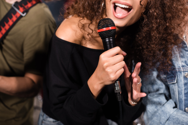 Cropped view of curly female vocalist with microphone singing near rock band musicians on blurred background - Photo, Image