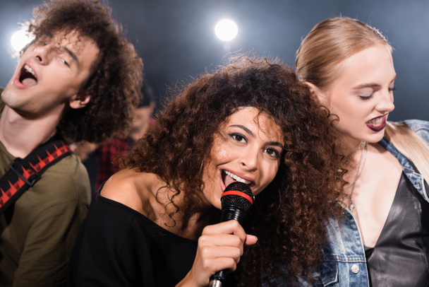 Mikrofonlu mutlu bir rock grubunun vokalisti arka planda bulanık arka planda çalan müzisyenlerin yanında şarkı söylüyor. - Fotoğraf, Görsel
