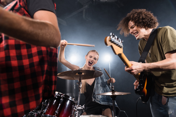 KYIV, UKRAINE - AUGUST 25, 2020: Blonde woman with drumsticks screaming while sitting at drum kit near guitarist with backlit and blurred man on foreground - Foto, afbeelding