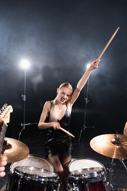 Blonde woman with outstretched hand winking while sitting near drum kit with backlit on blurred foreground - Photo, Image