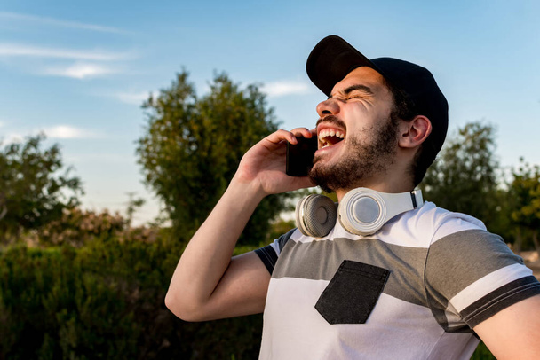 A young male with headphones and a cap laughing happily while talking with a phone in a park - Zdjęcie, obraz