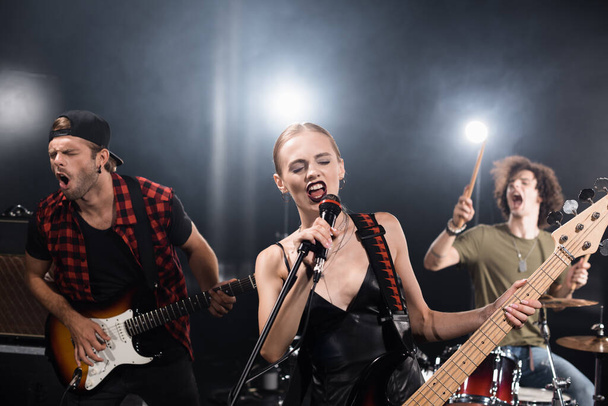 KIEW, UKRAINE - 25. AUGUST 2020: Blonder Sänger mit E-Gitarre singt im Mikrofon neben Gitarrist und Schlagzeuger mit Hintergrundbeleuchtung auf verschwommenem Hintergrund - Foto, Bild