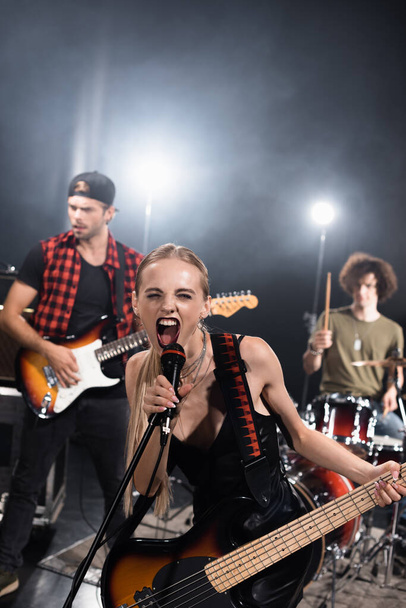 KYIV, UKRAINE - AUGUST 25, 2020: Female vocalist of rock band with electric guitar screaming in microphone with backlit and blurred musicians on background - Foto, Imagem
