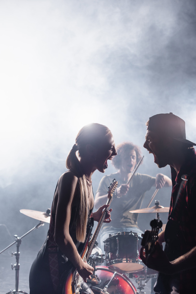 KYIV, UKRAINE - AUGUST 25, 2020: Rock band singers with bass guitars shouting and looking at each other with smoke and blurred drummer on background - Foto, afbeelding