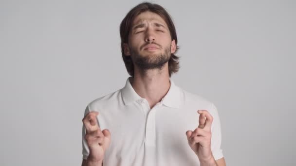 Νεαρός συναισθηματικός άντρας με σταυρωμένα δάχτυλα προσεύχεται πάνω από γκρι φόντο. Παρακαλώ εκφράστε - Πλάνα, βίντεο