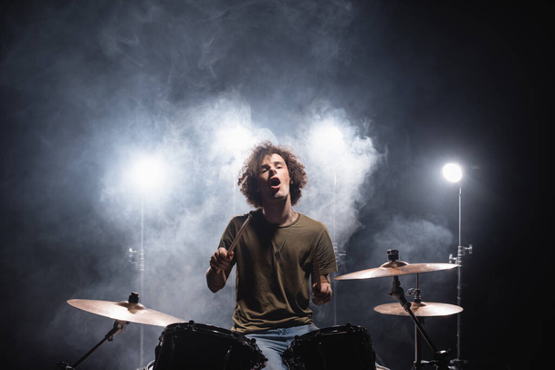 Opgewonden krullende drummer met open mond zittend aan drumstel met rook en achtergrondverlichting op zwart  - Foto, afbeelding