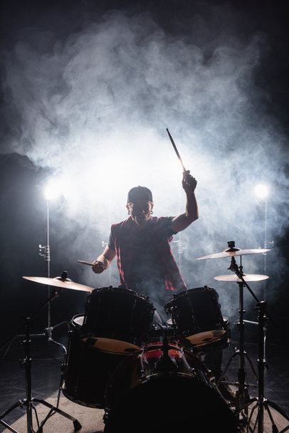 Человек играет на барабанах, сидя за барабанной установкой с подсветкой и дымом на заднем плане - Фото, изображение