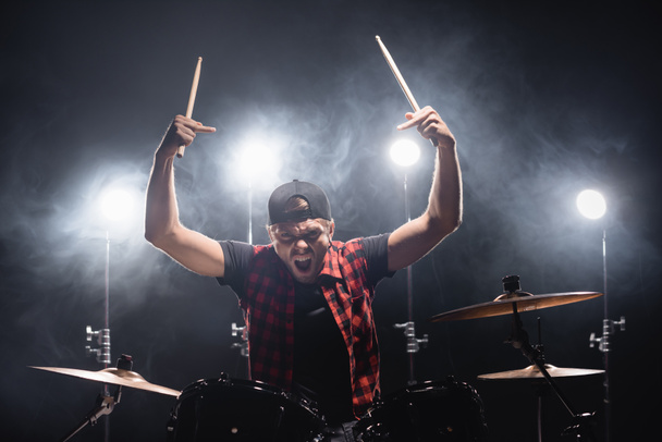 Aggressive drummer with middle fingers looking at camera and holding drumsticks near drum kit with backlit on background - Foto, imagen