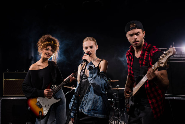 KYIV, UKRAINE - AUGUST 25, 2020: Female singer of rock band standing near musicians with electric guitars on black  - Foto, imagen