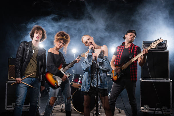 KYIV, UKRAINE - AUGUST 25, 2020: vocalist of rock band singing near musicians with bass guitars and drumsticks with smoke on black - Foto, imagen