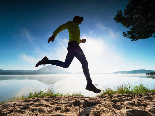 Un uomo caucasico che si allena sulla spiaggia domattina. Fitness runner in jersey giallo e collant neri entro la mattina correre all'aperto al lago. Daybreak paesaggio marino - Foto, immagini
