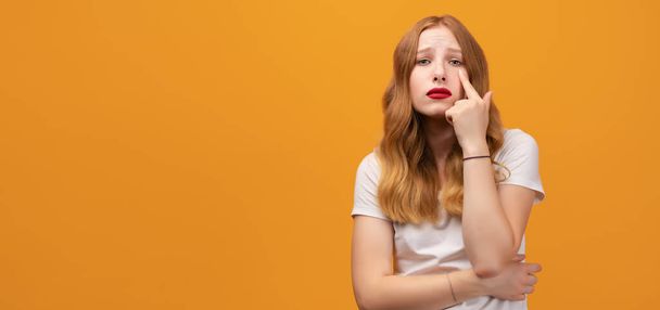 Pretty girl with wavy redhead, wearing white t-shirt pointing to the eye watching you gesture, suspicious expression. Isolated on yellow background. Copy space for your text - Foto, Imagen
