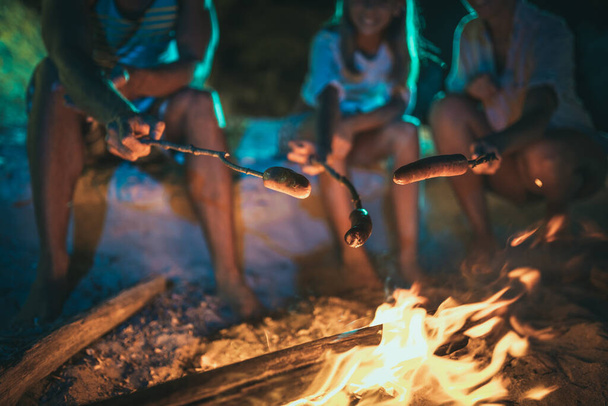 Una famiglia felice è seduta sulla spiaggia sabbiosa vicino al fuoco la sera. Stanno friggendo salsicce su bastone. - Foto, immagini
