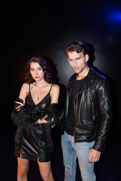 Sexy brunette woman with crossed arms standing near man in leather jacket with backlit on black - Zdjęcie, obraz