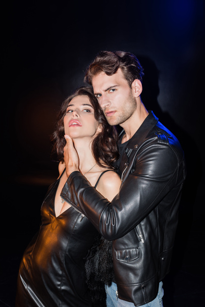 Confident man in leather jacket touching girlfriend neck, while looking at camera on black - Φωτογραφία, εικόνα