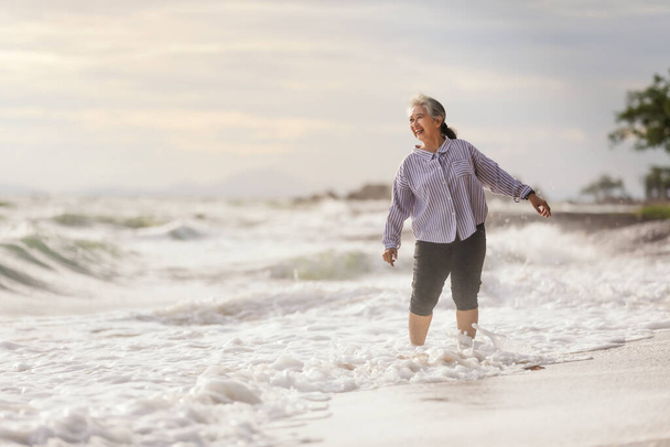 Yaşlı Asya 'lı kadın sahilde çıplak ayakla dans ediyor, dalgalar sahile vururken mutlu bir şekilde gülümsüyor. - Fotoğraf, Görsel