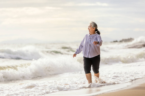 Старша Азія танцює на пляжі босоніж, Посміхнися з щасливо, коли хвиля в морі поспішає до берега - Фото, зображення