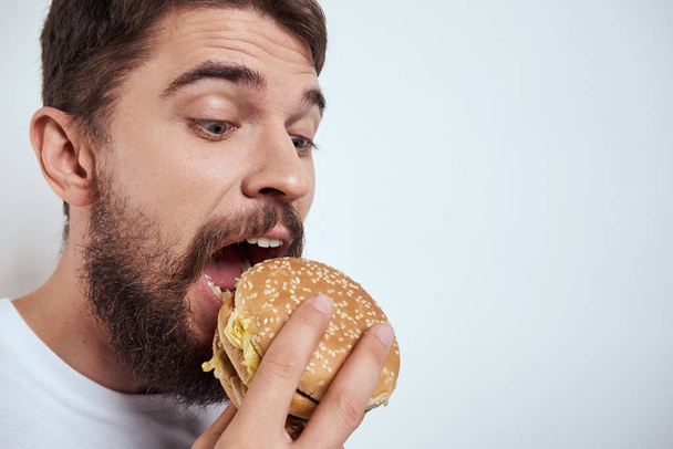Een man die een hamburger eet op een lichte achtergrond in een wit T-shirt cropped view close-up honger fast food - Foto, afbeelding