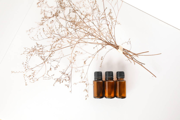 A closeup of small bottles of amber essential oil with dried plants on a white surface - Foto, Imagen