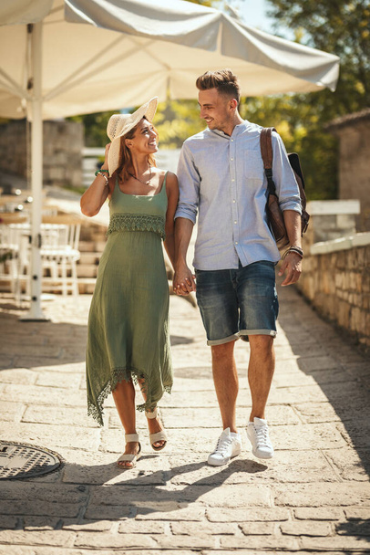 Una hermosa pareja joven se está divirtiendo mientras camina por una ciudad mediterránea. Están disfrutando en verano de un día soleado, tomándose de la mano y sonriendo. - Foto, Imagen