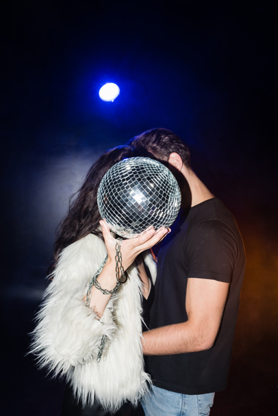 Curly woman in white jacket holding disco ball, while embracing with boyfriend with backlit on black - Photo, image