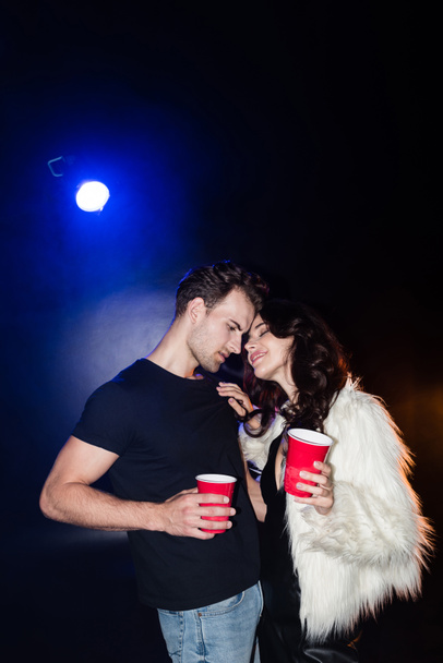 Smiling woman with closed eyes touching boyfriend shirt, while holding plastic cup with backlit on black - Fotoğraf, Görsel