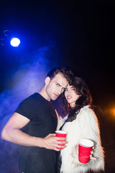 Irritated young adult couple looking at camera, holding plastic cups, while embracing with smoke and backlit on black - Fotografie, Obrázek
