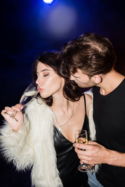 Passionate man looking at sexy brunette woman with closed eyes drinking champagne with backlit on black - Photo, Image