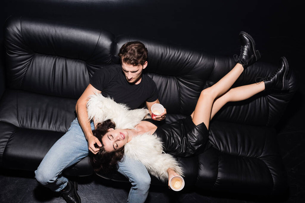 High angle view of woman with legs in air holding plastic cup, while lying on man in casual clothing on sofa in nightclub - Фото, зображення