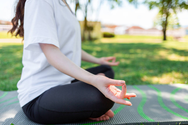 Du yoga. Une femme en tenue de sport médite les mains pliées sur ses genoux, assise sur un tapis, dans un parc sur l'herbe. Vue latérale. Gros plan de la main. Concept de mode de vie sain et d'harmonie. - Photo, image
