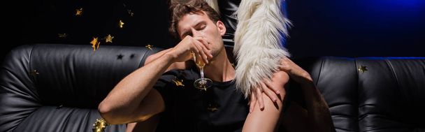 Macho férfi pezsgőt iszik, kéz a kézben női láb ül a kanapén, míg konfetti esik nightclub, banner - Fotó, kép