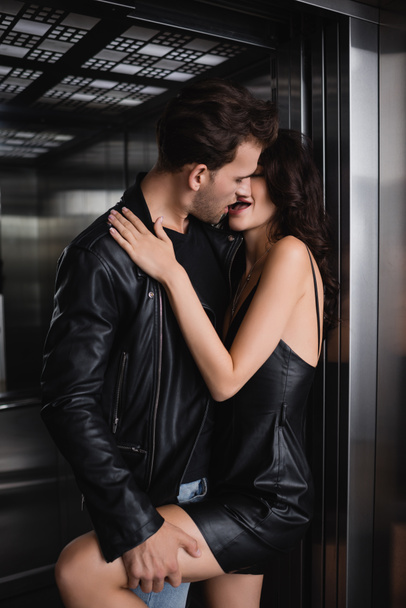 Homme en veste en cuir embrasser et tenir jambe de femme séduisante en robe noire dans l'ascenseur - Photo, image