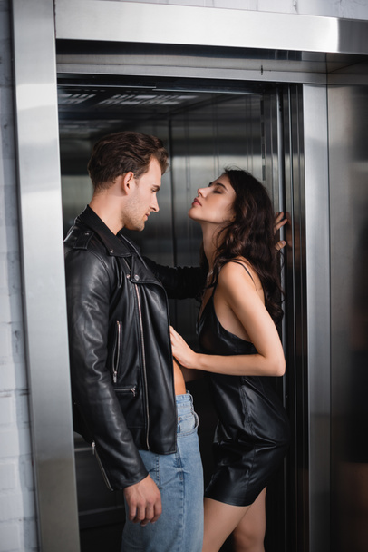 Страстная кудрявая женщина с закрытыми глазами раздевает мужчину в кожаной куртке в лифте - Фото, изображение