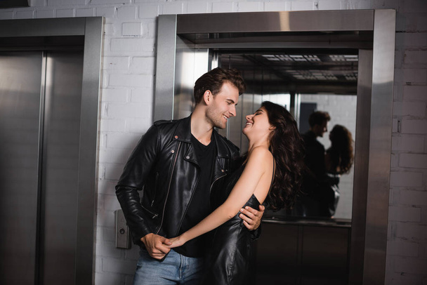 Улыбающийся мужчина в черной куртке обнимает и держит за руку веселую кудрявую женщину в кожаном платье у входа в лифт - Фото, изображение