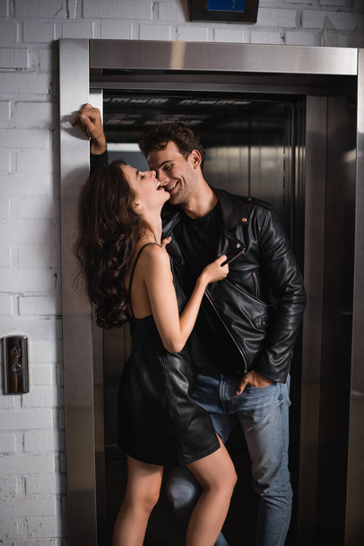 Smiling man with hand in pocket looking at laughing girlfriend touching jacket, while leaning on elevator entrance - Photo, Image