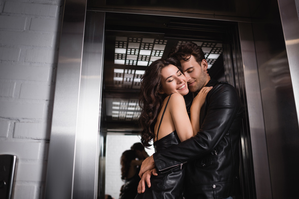 Улыбающийся мужчина с закрытыми глазами обнимает счастливую брюнетку у входа в лифт - Фото, изображение