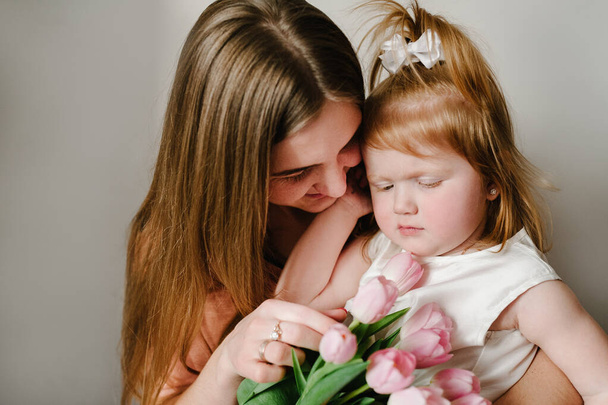 The mother holds on his hands little girl on light background indoor. Portrait of mom with flowers and daughter sitting in home. The concept of happy family holiday. woman's day. Mother's Day. - Fotó, kép