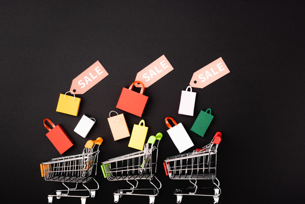 Vista superior de carritos de juguete y bolsas de compras con etiquetas de precios y letras de venta sobre fondo negro  - Foto, imagen