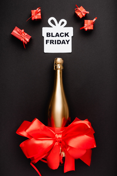 Top view arany pezsgős üveg piros íj és papír ajándék fekete péntek betűkkel fekete háttér  - Fotó, kép