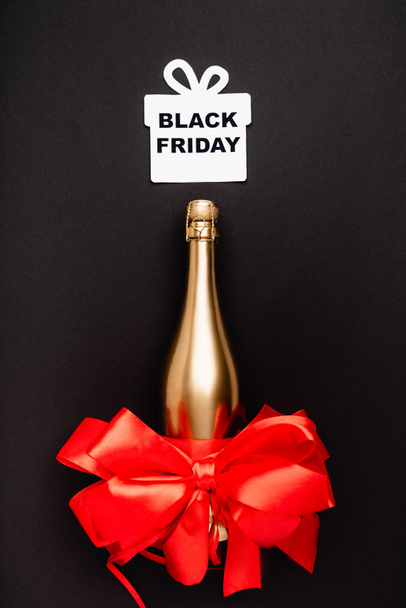 Widok z góry złotej butelki szampana, czerwony łuk i papier prezent z czarnym piątkowym napisem na czarnym tle  - Zdjęcie, obraz