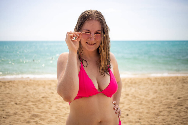 junges verspieltes kaukasisches Mädchen lächelnd, flirtend und in die Kamera blickend, bekleidet mit rosa Bikini und Brillenherzen am Strand - Foto, Bild