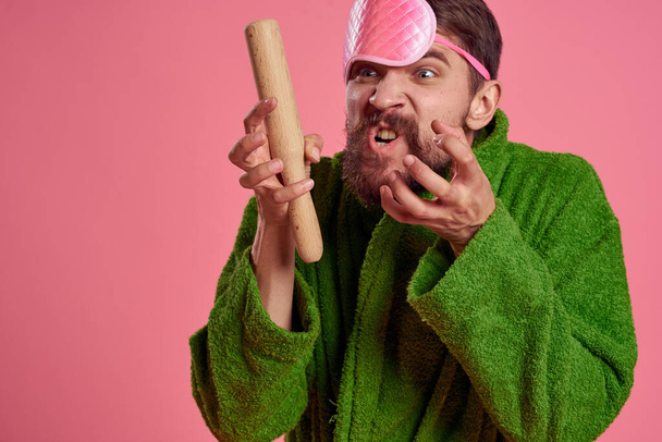 Retrato de un hombre con una máscara de sueño rosa y un rodillo de madera emociones modelo de irritabilidad bata verde - Foto, Imagen