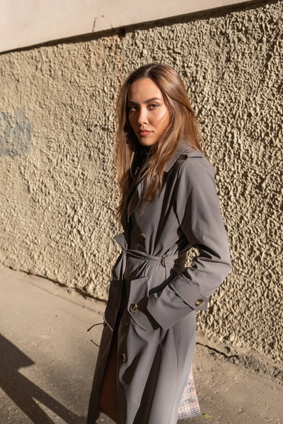 attraktives junges asiatisches Model im grauen Trenchcoat, das an der Wand im Freien steht - Foto, Bild