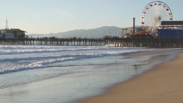 Vagues océaniques et plage de sable californienne, roue ferris classique dans un parc d'attractions sur jetée à Santa Monica Pacific Ocean Resort. Vue emblématique de l'été, symbole de Los Angeles, CA USA. Concept de voyage - Séquence, vidéo