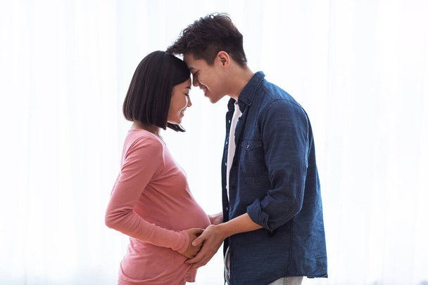 窓の近くに立つアジアの夫妊娠中の妻に触れる腹屋内 - 写真・画像
